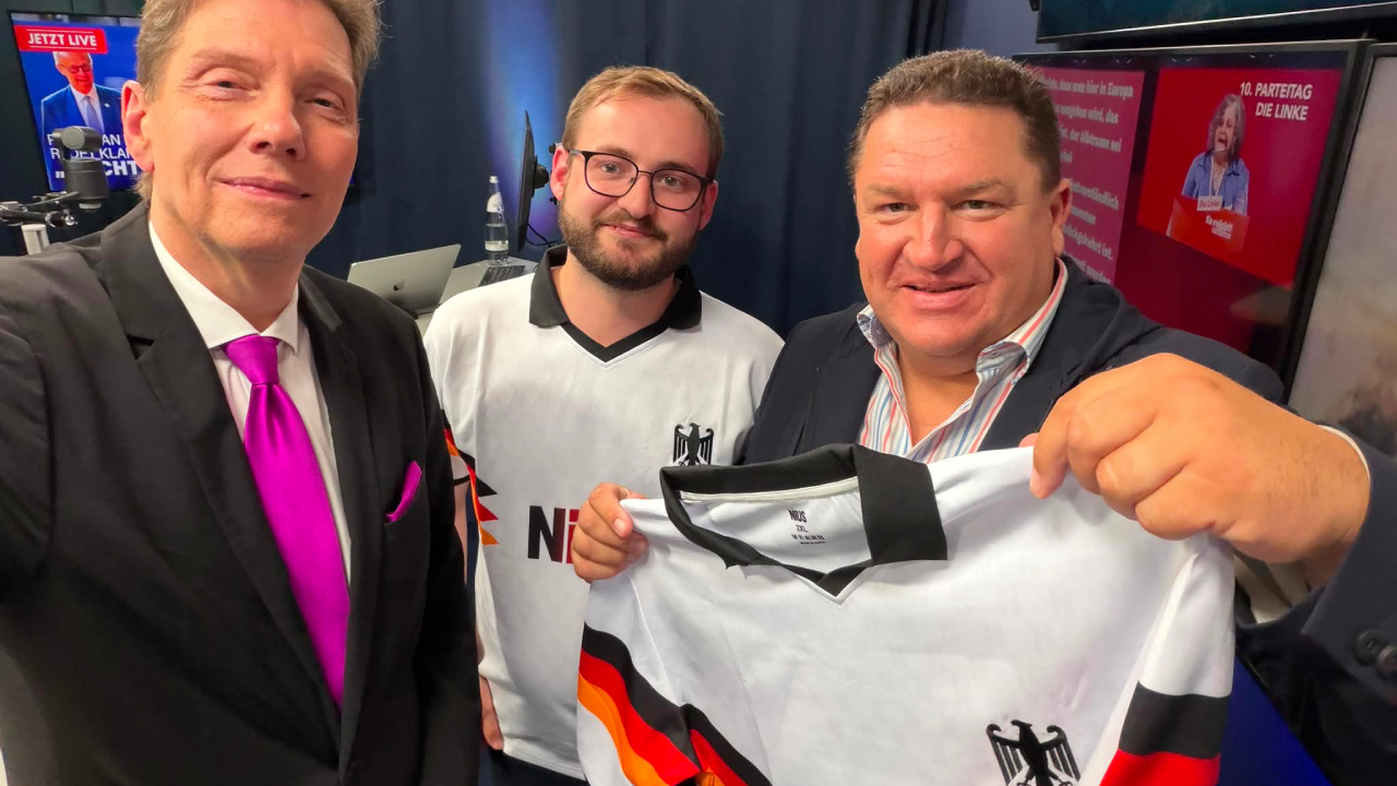 Von links nach rechts: Moderator Andreas Dorfmann, NIUS-Reporter Philippe Fischer und Marcus Held im NIUS Studio. Marcus Held präsentiert ein NIUS-Nationaltrikot im Rahmen einer Sendung zu aktuellen politischen Themen und Reformdebatten in Deutschland.